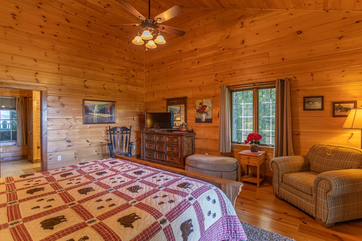 This serene bedroom features wooden walls, a cozy bed with a quilt, an armchair for reading, and a window with nature views. The calm atmosphere and separate layout make it a great fit for representing a lower queen bedroom or the upper loft queen suite.