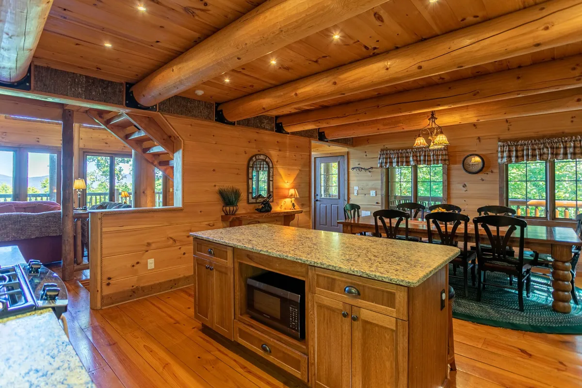 This broad interior view highlights the gourmet kitchen with a granite island, seamlessly flowing into the dining area. Large windows frame mountain vistas, and exposed wooden beams enhance the rustic, crafted feel. The kitchen is equipped for cooking and entertaining, with ample space for guests to gather.