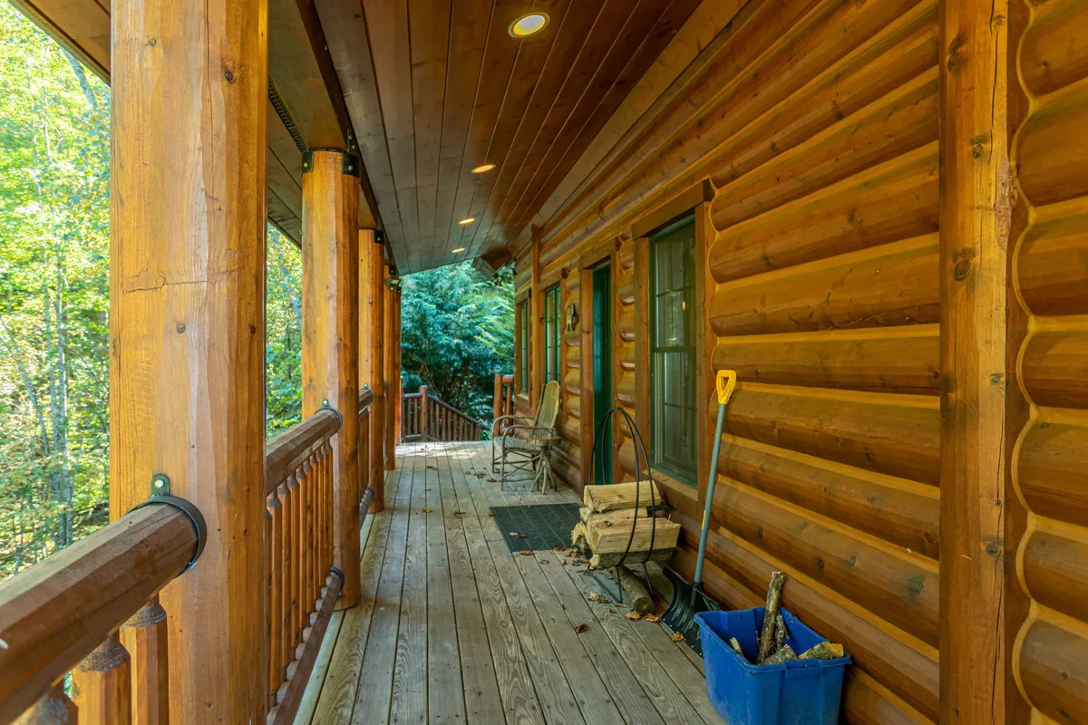 The image shows the exterior of a log cabin, with a wooden porch featuring rocking chairs and a rustic blue container for firewood. The setting is surrounded by trees, emphasizing a cozy yet adventurous atmosphere ideal for relaxing after a day outdoors.