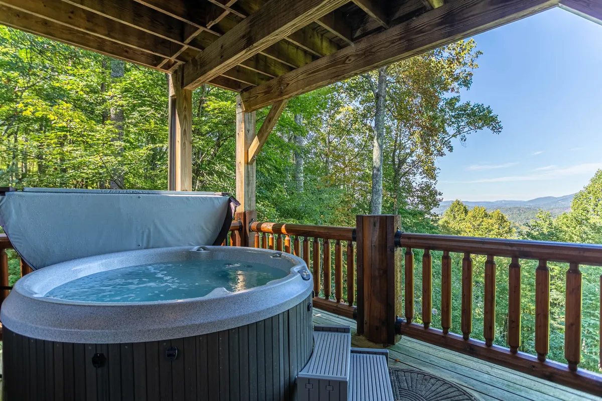 An inviting exterior shot of a private covered deck featuring a bubbling hot tub, wooden railing, and panoramic mountain views. The mood is tranquil and ideal for stargazing or soaking after a day of adventure.