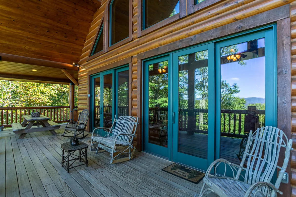 This image features the log cabin structure with expansive wooden decks, large glass doors, rocking chairs, and a picnic table set against a scenic backdrop. The decks wrap around the cabin, inviting guests to relax while taking in the natural surroundings.