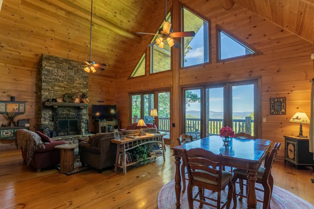 This image captures the core of the Great Room, showcasing a majestic stone fireplace, expansive windows flooding the space with natural light and mountain views, a large wooden dining table, and a plush seating area arranged for gatherings. The knotty pine paneling and artisan-crafted woodwork are visible, underscoring the room’s rustic elegance and welcoming ambiance.