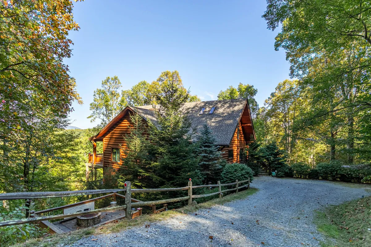 The image showcases a charming log cabin nestled among tall trees, with a welcoming gravel driveway and rustic wooden fence framing the property. The tranquil setting hints at scenic mountain surroundings, and the spacious porch invites guests to unwind and enjoy nature.