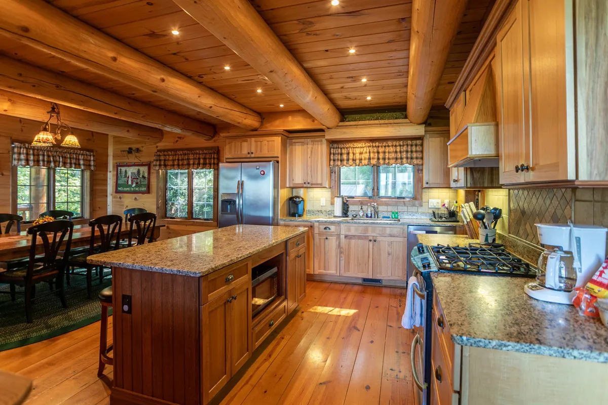 This image offers a closer look at the kitchen and part of the dining area, with honed granite countertops, custom wooden cabinetry, and a large dining table bathed in natural light from the windows. Bar stools and stainless appliances are also visible, inviting both food preparation and social interaction.