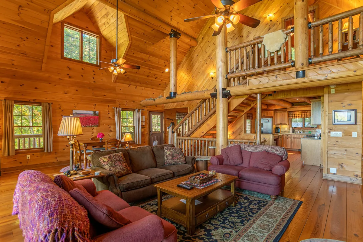 A welcoming living room scene featuring rustic wooden beams, comfortable sofas, a central coffee table, and a staircase, all bathed in natural light. Large windows suggest a connection to the outdoors, with a layout that invites relaxation and socializing after a day of mountain exploration.