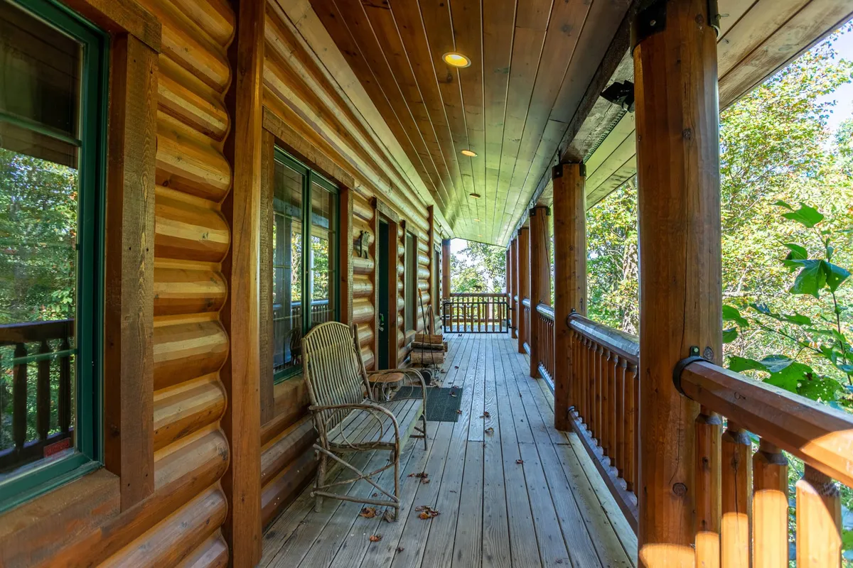 A wide, welcoming porch lined with classic wooden rocking chairs offers views of the surrounding green landscape. The log walls and wooden flooring create a cozy, rustic feel, inviting guests to relax, converse, or soak in the mountain atmosphere. The railing frames the porch, giving a sense of both openness and intimacy with nature.