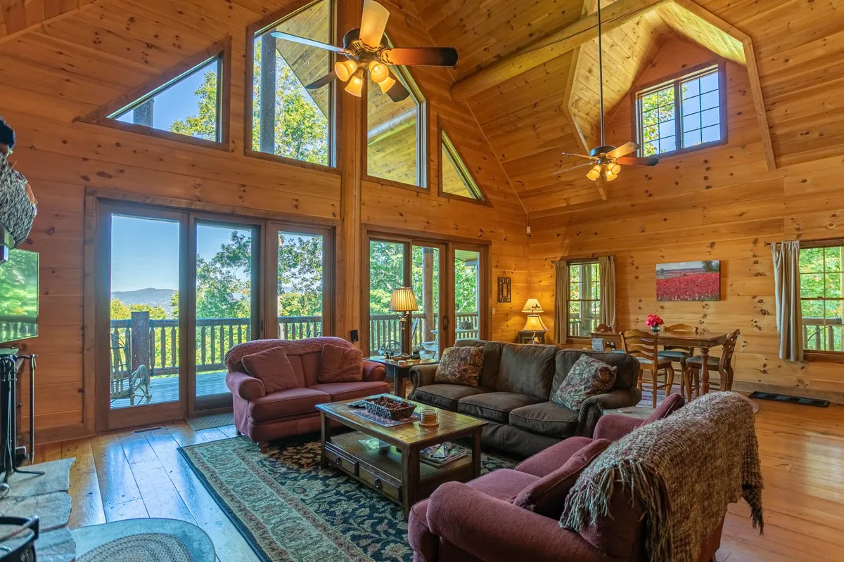 This image displays a spacious interior with large windows framing mountain views, exposed wooden beams, and a comfortable seating and dining area. Natural light pours in, suggesting an inviting spot to relax with coffee in the morning or unwind after a day exploring.